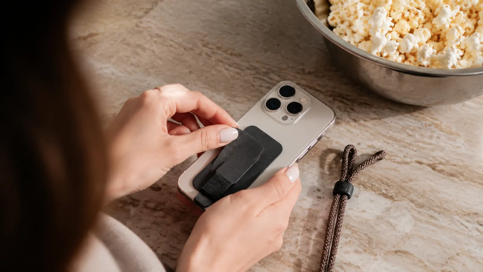 Close-up of hands pressing a small adhesive tab to the back of a phone on a kitchen counter, a woven non-metal cord lying beside it