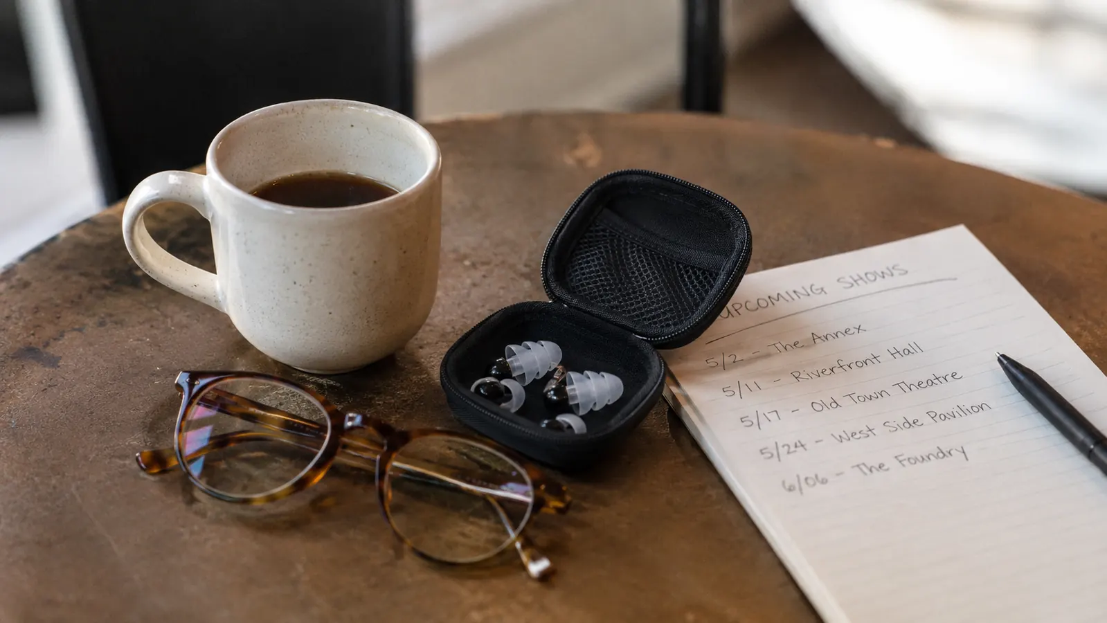 A small earplug carry case resting on a table beside a handwritten list of shows and a pair of reading glasses