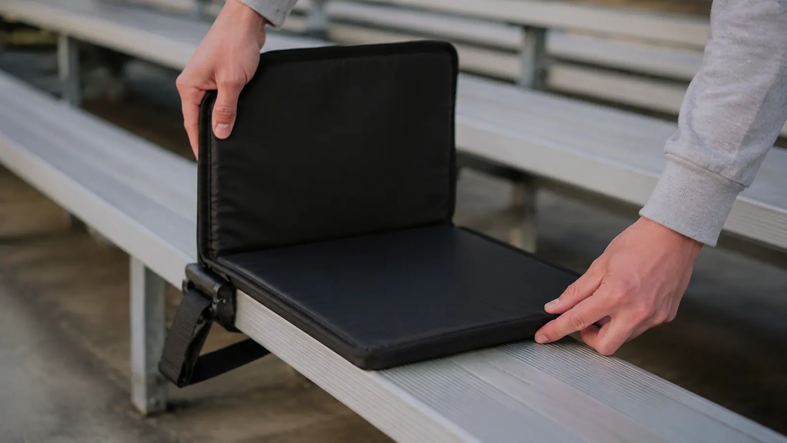 Two hands securing a folding spectator seat onto the plank of an outdoor bleacher bench