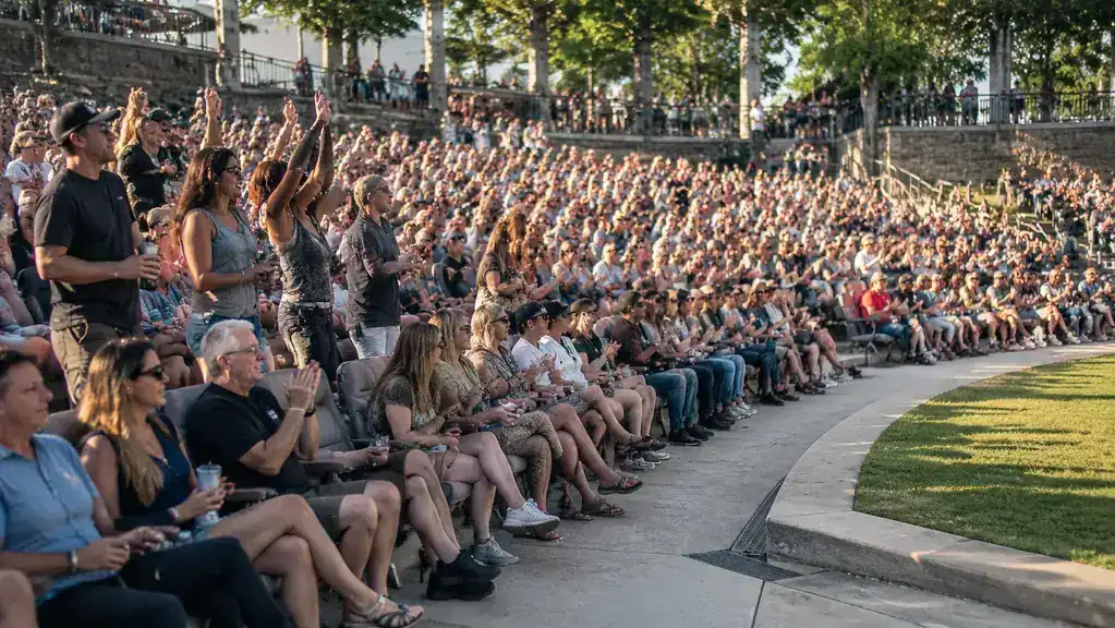 A daytime festival crowd filling an outdoor lawn and bench seating, many people sitting in padded folding stadium chairs, warm afternoon light, hands raised toward the stage